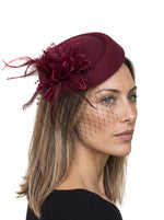 A woman in profile wearing a burgundy fascinator hat with a matching feather and flower embellishment, black netting veil, and blonde hair against a white background.